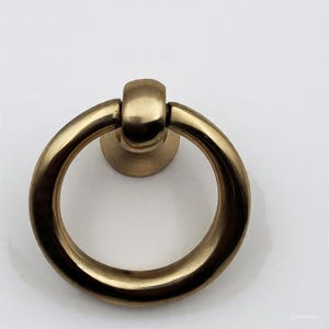 May include: A close-up of a brass ring pull with a circular handle and a round base. The hardware has a warm, golden hue and is designed for furniture. The ring is attached to a small, circular base.