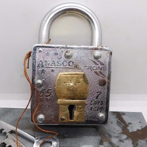 May include: A vintage silver padlock with a brass-colored star-shaped emblem. Two silver keys are connected by a brown string. The padlock is on a gray and gold marbled surface. The padlock has the word 'ATLASCO' and the numbers '55' and '7' engraved on it.