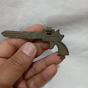 May include: A vintage key shaped like a pistol, featuring a detailed handle and barrel. The key is made of a dark, aged metal, with visible wear and tear. The key is held in a hand against a white background.