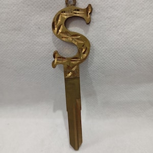 May include: A vintage brass key with an ornate, stylized "S" design. The key features a decorative loop at the top and a detailed pattern etched into the letter. The key's blade is rectangular and tapers to a point.