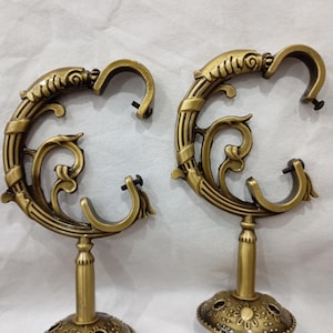 May include: Two antique bronze curtain rod holders. Each holder features an ornate, swirling design with a curved hook and a decorative base. The holders are made of metal and have a vintage aesthetic.