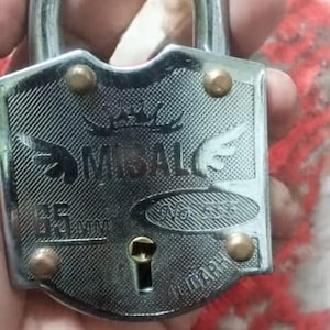 Pode incluir: Um cadeado prateado com uma alça curva. A fechadura tem a marca "MISAL" e a medida "65 mm" impressa na frente. Quatro rebites de latão prendem a placa frontal.