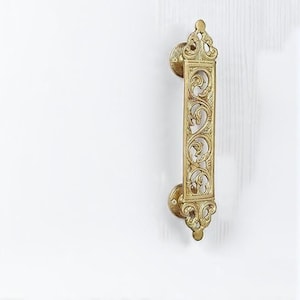 May include: Ornate brass door pull with a detailed, openwork design. The handle features scrolling patterns and decorative end pieces, mounted on a white surface. A vintage-style hardware element.
