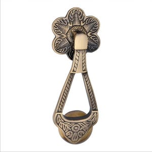 Op de afbeelding: Een antieke deurklopper in messingkleur met een bloemmotief. De klopper heeft een bloemvormige montageplaat, een druppelvormige handgreep en een decoratieve bal aan de onderkant. Het ontwerp bevat ingewikkelde blad- en bloempatronen.