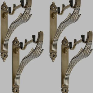 May include: Four antique brass curtain rod holders. Each holder features a decorative, fish-scale design and a double hook for hanging curtains. The holders have a vertical, rectangular backplate with an ornate top and bottom.