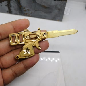Puede incluir: Una llave dorada con forma de pistola. La llave tiene un diseño detallado, que incluye un gatillo, un cañón y elementos florales decorativos. La llave se sostiene en una mano sobre un fondo blanco.