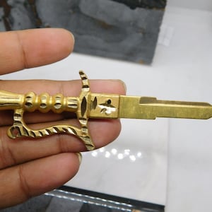 Puede incluir: Una llave dorada con un mango decorativo en forma de espada. La llave tiene un diseño detallado en el mango y un extremo en forma de hoja. La llave se sostiene en una mano.
