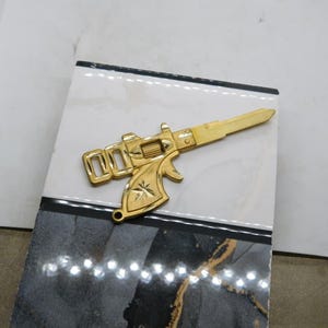 Pu&ograve; includere: Una chiave color oro a forma di pistola, appoggiata su una piastrella decorativa. La chiave presenta un design dettagliato, tra cui un emblema a forma di stella. La piastrella ha un motivo marmorizzato bianco e nero.