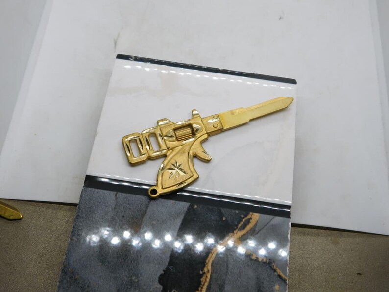 Pu&ograve; includere: Una chiave color oro a forma di pistola, appoggiata su una piastrella decorativa. La chiave presenta un design dettagliato, tra cui un emblema a forma di stella. La piastrella ha un motivo marmorizzato bianco e nero.
