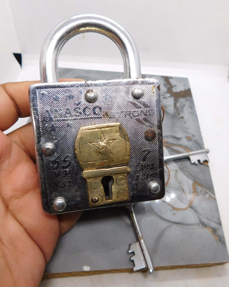 Puede incluir: Un candado plateado vintage con un emblema en forma de estrella de color lat&oacute;n. El candado tiene el texto "NASCO IRON" y viene con dos llaves plateadas. El candado se sostiene en una mano.