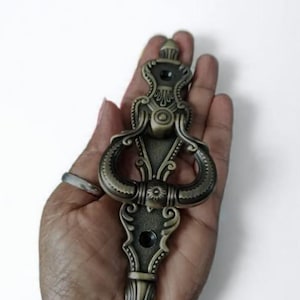 Op de afbeelding: Een antieke deurklopper in bronskleur met sierlijke details. De klopper heeft een decoratieve bovenkant, gebogen handvatten en een gedetailleerd onderste gedeelte. Het ontwerp bevat bloemen- en krulwerk elementen, wat een vintage uitstraling creëert.