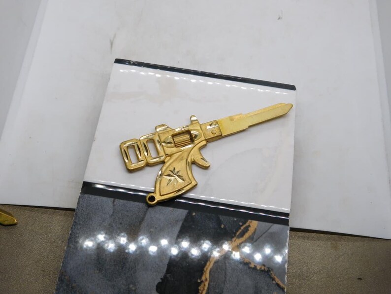Pu&ograve; includere: Una chiave color oro a forma di pistola, con un design dettagliato. La chiave &egrave; appoggiata su una superficie decorativa con motivi neri, bianchi e grigi. La canna e l'impugnatura della chiave sono ben visibili.