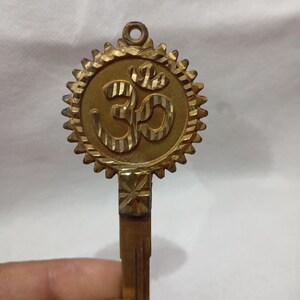 May include: A gold-colored metal pendant with the Om symbol in the center. The pendant has a circular design with a sunburst border and a key-like shape below. The Om symbol is etched with a textured pattern.