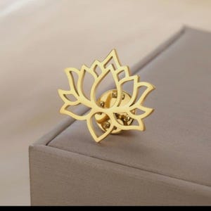 May include: A gold-colored lotus flower pin. The pin features a detailed cutout design of the flower's petals. The pin is made of a shiny, metallic material and is set against a neutral background.