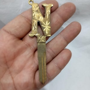 May include: A vintage brass key with the letter "N" as the key's head. The letter is decorated with floral patterns. The key has a loop at the top for attachment and a blade at the bottom. The key is held in a hand.