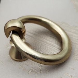 May include: A close-up of a brass ring pull with a circular handle and a decorative base. The hardware is a warm gold color and appears to be made of metal. The ring is attached to a small, rounded base.