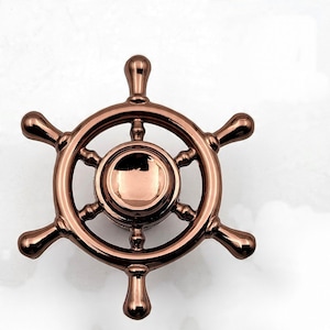Puede incluir: Un timón de color bronce, con seis radios y un cubo central circular. El volante tiene un acabado metálico brillante y se encuentra sobre un fondo blanco. Este artículo decorativo de temática náutica es adecuado para la decoración del hogar.