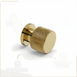 Puede incluir: Un pomo de armario dorado con un agarre texturizado con patrón de diamantes. El pomo tiene una base plana y circular y un extremo liso y redondeado. El herraje está diseñado para muebles y decoración del hogar.