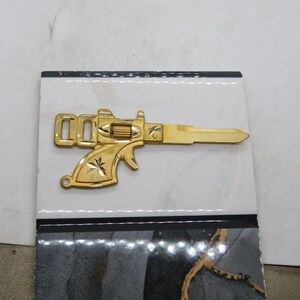 Pu&ograve; includere: Una chiave color oro a forma di pistola, con dettagli intricati sull'impugnatura e sulla canna. La chiave &egrave; posta su uno sfondo a motivi bianchi e neri, creando un contrasto sorprendente. Il design della chiave ricorda un'arma da fuoco vintage.