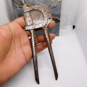 May include: A vintage, rusty metal nutcracker with a curved handle and two long, pointed levers. The nutcracker has a weathered appearance, suggesting age and use. The design is simple and functional, with a focus on durability.