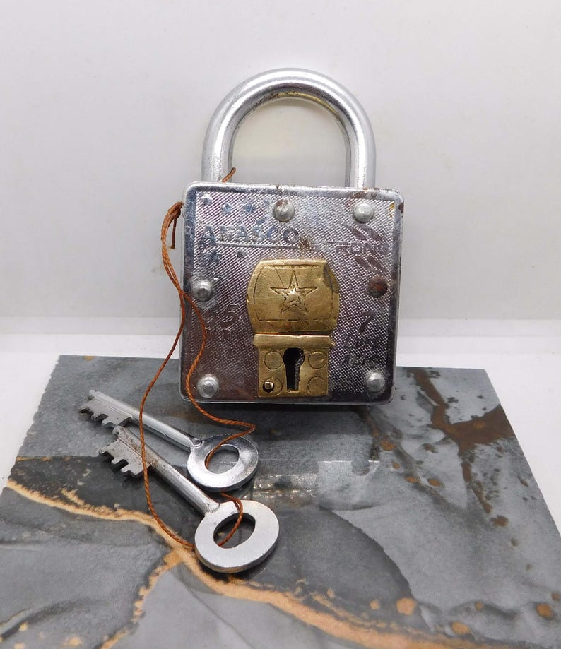 May include: A vintage silver padlock with a brass-colored star-shaped emblem. Two silver keys are connected by a brown string. The padlock is on a gray and gold marbled surface. The padlock has the word 'ATLASCO' and the numbers '55' and '7' engraved on it.