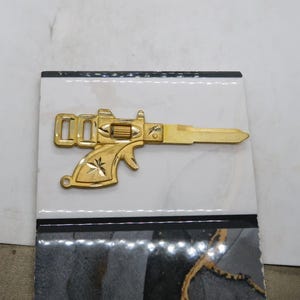 Pu&ograve; includere: Una chiave color oro a forma di pistola, con dettagli intricati sull'impugnatura e sulla canna. La chiave &egrave; posta su uno sfondo a motivi bianchi e neri, creando un contrasto sorprendente. Il design della chiave ricorda un'arma da fuoco vintage.