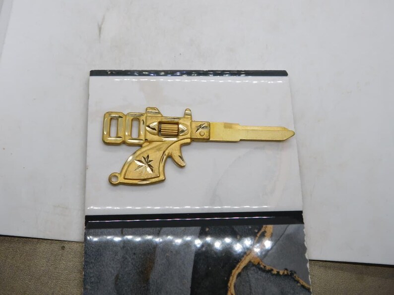 Pu&ograve; includere: Una chiave color oro a forma di pistola, con dettagli intricati sull'impugnatura e sulla canna. La chiave &egrave; posta su uno sfondo a motivi bianchi e neri, creando un contrasto sorprendente. Il design della chiave ricorda un'arma da fuoco vintage.