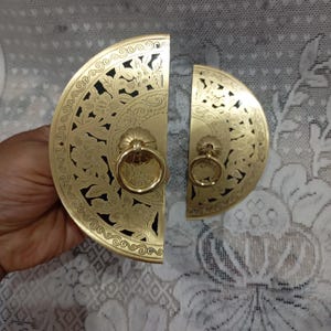 May include: Two semi-circular brass cabinet pulls featuring intricate floral cut-out patterns and central ring handles. The metal hardware displays a polished gold-tone finish with detailed engravings along the curved edges and a solid flat base.