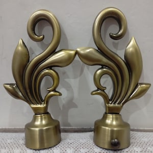 May include: A pair of antique brass-colored curtain rod finials featuring a symmetrical, ornate scrollwork design with leaf-like accents. Each finial has a cylindrical base with a small screw mechanism visible on the side for secure attachment.