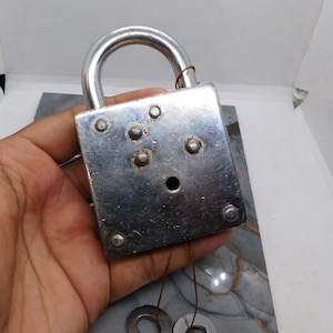 Puede incluir: Un candado plateado vintage con un asa curva y dos llaves unidas por finos cordones. El candado tiene un cuerpo rectangular con varios remaches y un ojo de cerradura. Las llaves tienen mangos circulares.
