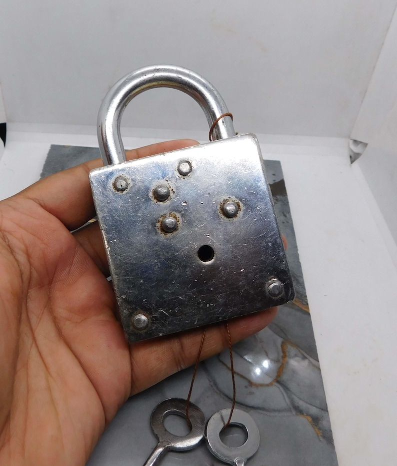 Puede incluir: Un candado plateado vintage con un asa curva y dos llaves unidas por finos cordones. El candado tiene un cuerpo rectangular con varios remaches y un ojo de cerradura. Las llaves tienen mangos circulares.
