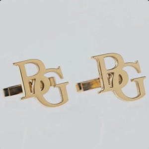Puede incluir: Un par de gemelos dorados. Cada gemelo presenta las letras "BG" en una fuente estilizada. Los gemelos tienen un poste rectangular para insertar en el puño. El fondo es de un color neutro.