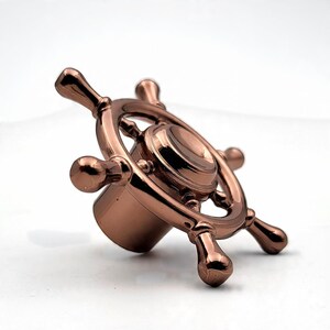 Peut inclure: Un bouchon de bouteille de couleur bronze en forme de gouvernail de navire. Le bouchon présente un design circulaire avec un disque central et six rayons. La finition métallique lui donne un aspect poli et réfléchissant.