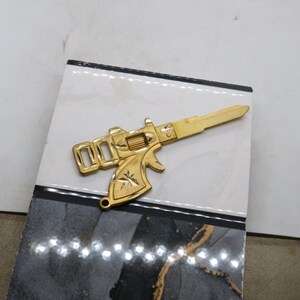Pu&ograve; includere: Una chiave color oro a forma di pistola, appoggiata su una superficie decorativa. La chiave presenta un design dettagliato, tra cui un grilletto, una canna e un'impugnatura. La superficie &egrave; un mix di bianco, nero e grigio, con finitura lucida. Un accessorio unico.