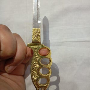 Puede incluir: Una llave decorativa con un mango de color latón diseñado con forma de nudillera. El mango presenta un patrón floral grabado y tres orificios para los dedos. La hoja de la llave es de color plateado y tiene una punta. Una piedra rosa está engastada en el mango.