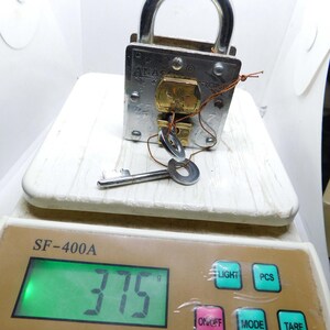 Puede incluir: Un candado plateado con un asa curva y una llave insertada. El candado est&aacute; sobre una balanza blanca, que muestra un peso de 375 gramos. La balanza tiene botones para luz, PCS, modo y tara.