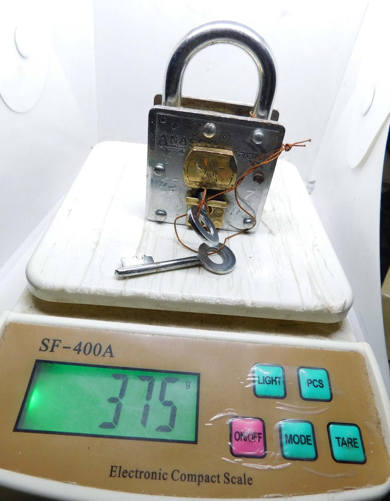 Puede incluir: Un candado plateado con un asa curva y una llave insertada. El candado est&aacute; sobre una balanza blanca, que muestra un peso de 375 gramos. La balanza tiene botones para luz, PCS, modo y tara.