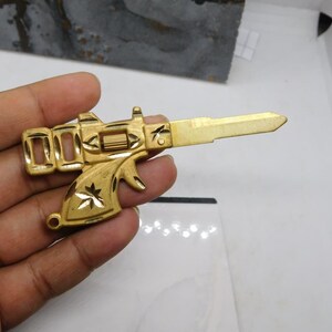 Pu&ograve; includere: Una chiave color oro a forma di pistola. La chiave presenta un design dettagliato, tra cui un grilletto, una canna e elementi floreali decorativi. La chiave &egrave; tenuta su uno sfondo bianco.