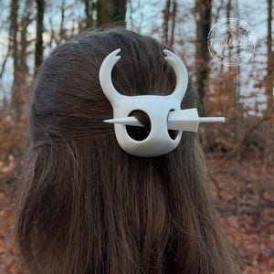 White 3D printed Hollow Knight hair clip worn in long brown hair, photographed in an autumn forest. The hair accessory features the iconic vessel mask design with curved horns and a circular opening, secured with a matching pin/stick. Shown from the back against a background of bare trees and fallen leaves.