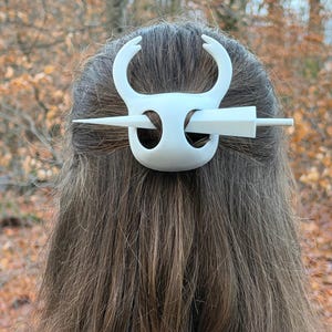 May include: White hair accessory featuring a stylized bull's head design. The hair clip has curved horns and a central bar that secures the hair. The accessory is made of a solid white material and is worn in the hair.