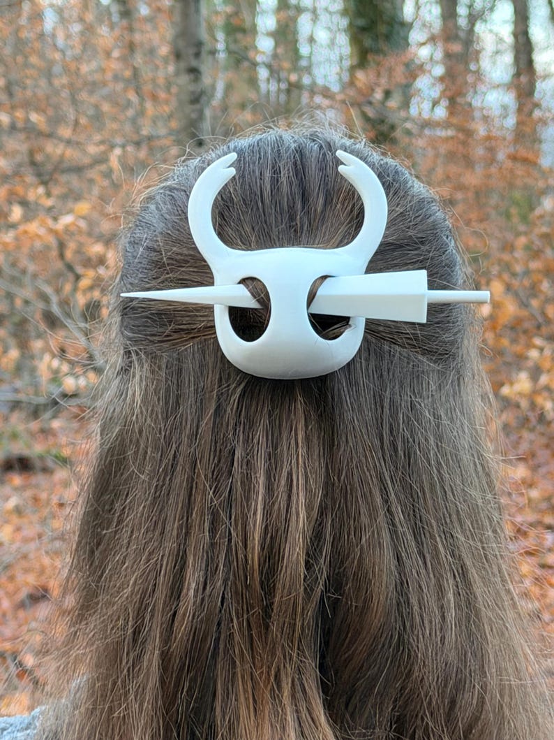 May include: White hair accessory featuring a stylized bull's head design. The hair clip has curved horns and a central bar that secures the hair. The accessory is made of a solid white material and is worn in the hair.