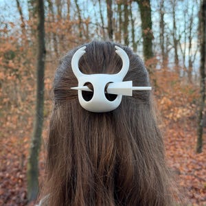 May include: A white hair accessory featuring a stylized design with curved horns and a central pin. The hair piece is set against a backdrop of brown hair and a blurred outdoor scene with trees.
