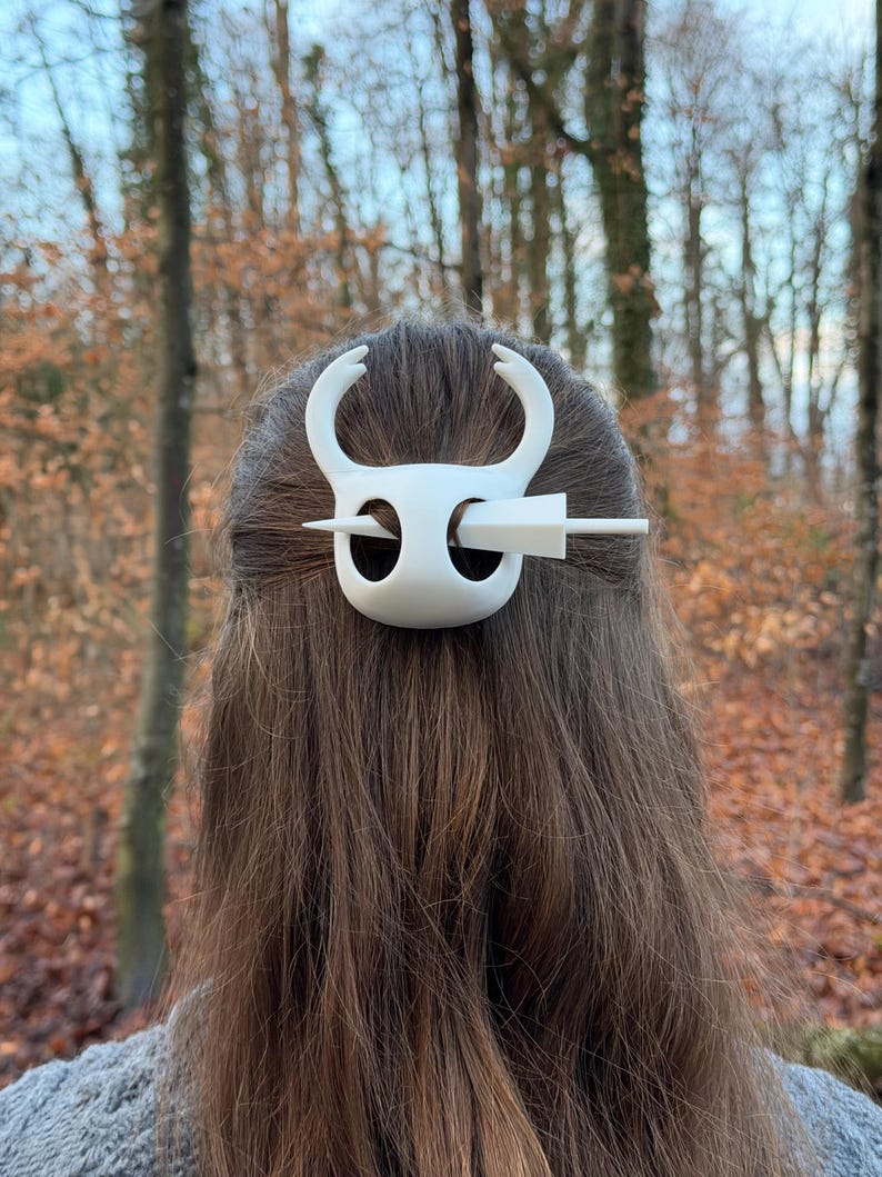 May include: A white hair accessory featuring a stylized design with curved horns and a central pin. The hair piece is set against a backdrop of brown hair and a blurred outdoor scene with trees.