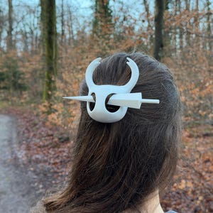 May include: A white hair accessory, featuring a stylized design with curved elements and a central pin. The accessory is worn in the hair, with the back of the head and hair visible. The background shows a blurred forest scene.