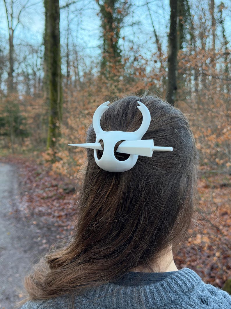 May include: A white hair accessory, featuring a stylized design with curved elements and a central pin. The accessory is worn in the hair, with the back of the head and hair visible. The background shows a blurred forest scene.