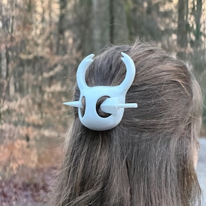 May include: A white hair clip, featuring a stylized design with curved horns and a central bar, is secured in brown hair. The clip's design is symmetrical and appears to be made of plastic. The background is blurred, with a natural outdoor setting.