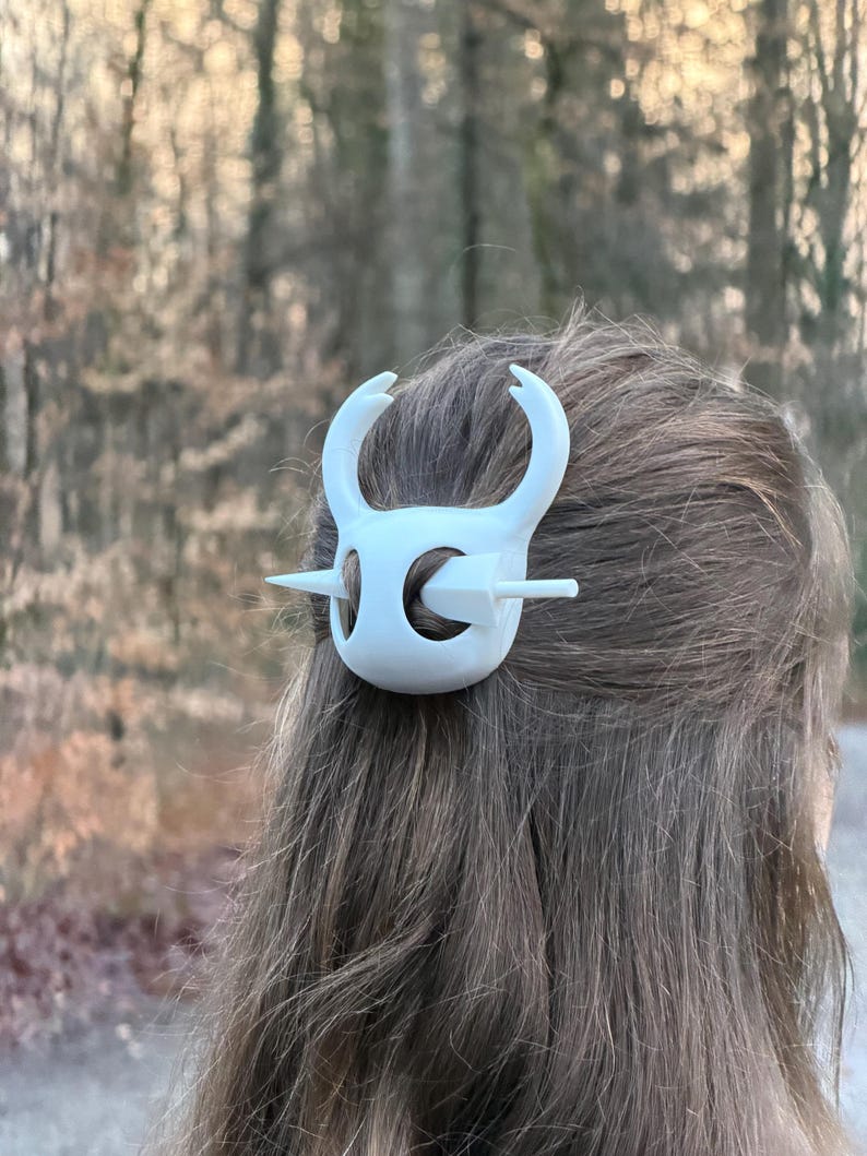 May include: A white hair clip, featuring a stylized design with curved horns and a central bar, is secured in brown hair. The clip's design is symmetrical and appears to be made of plastic. The background is blurred, with a natural outdoor setting.