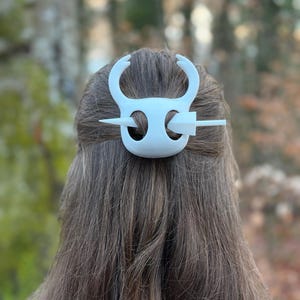 May include: White hair accessory with a decorative design, featuring curved elements and a horizontal pin. The accessory is set against a backdrop of brown hair and a blurred outdoor setting, suggesting a nature-inspired theme.