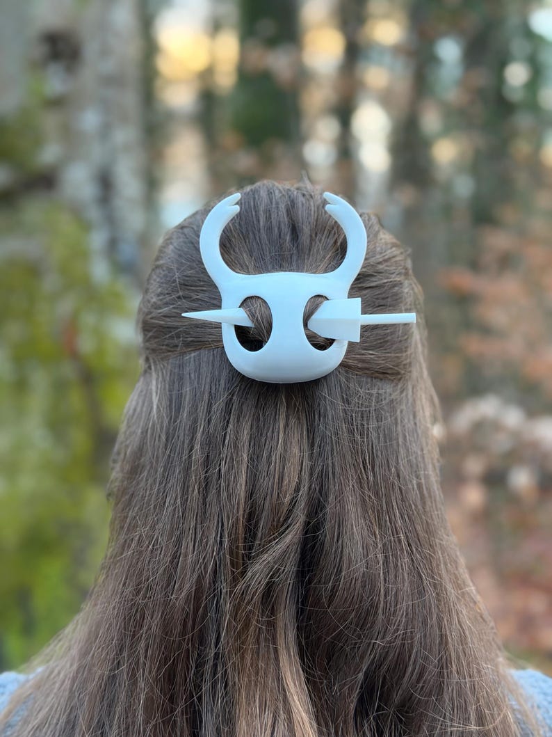 May include: White hair accessory with a decorative design, featuring curved elements and a horizontal pin. The accessory is set against a backdrop of brown hair and a blurred outdoor setting, suggesting a nature-inspired theme.