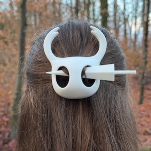 May include: A white hair clip, featuring a stylized design with curved horns and a central pin, is inserted into brown hair. The clip's design is reminiscent of a character from a video game. The background is a blurred view of a forest.